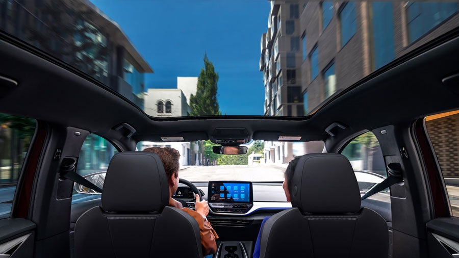 Backseat view looking forward between the driver and passenger seats into the cockpit area of an ID.4. We see a woman in the passenger seat and a man driving them down a city street lined with modern buildings. Through the panoramic glass roof a blue sky and the tops of buildings are seen